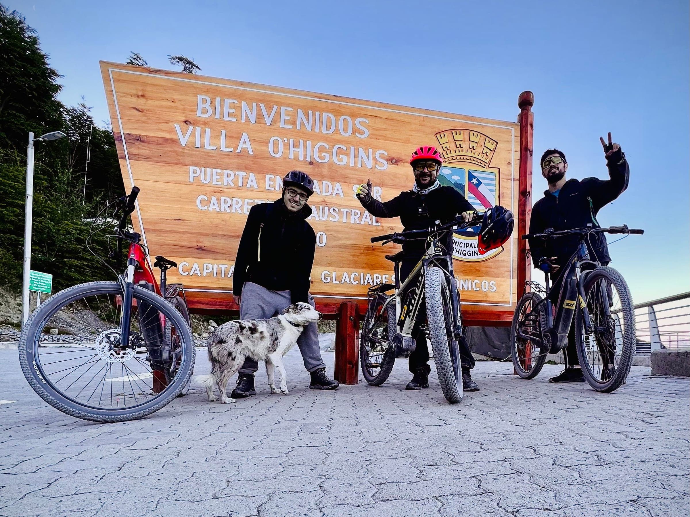 Tre ciclisti con le loro e-bike e un cane davanti al cartello di benvenuto di Villa O'Higgins