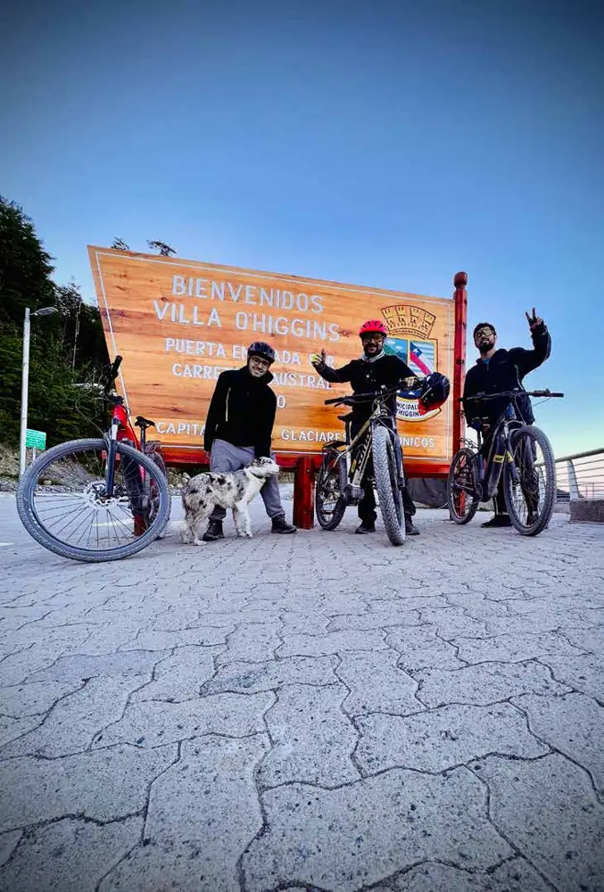 Tre ciclisti con le loro bici e un cane accanto al cartello di benvenuto di Villa O'Higgins
