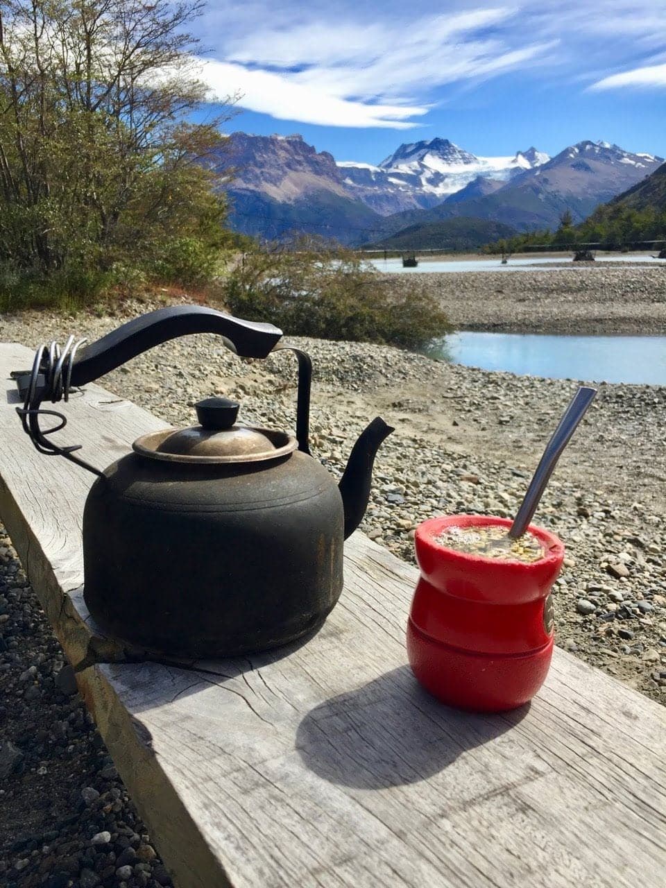 Un bollitore nero e un mate rosso su un tavolo di legno, davanti al lago e alla cordigliera